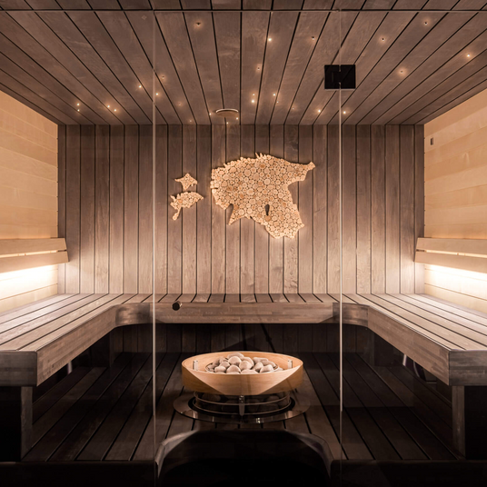 Modern sauna room with wooden benches, soft ambient lighting, and a bowl of stones in the center. A wooden map of Estonia is mounted on the back wall, illuminated by spotlights.
