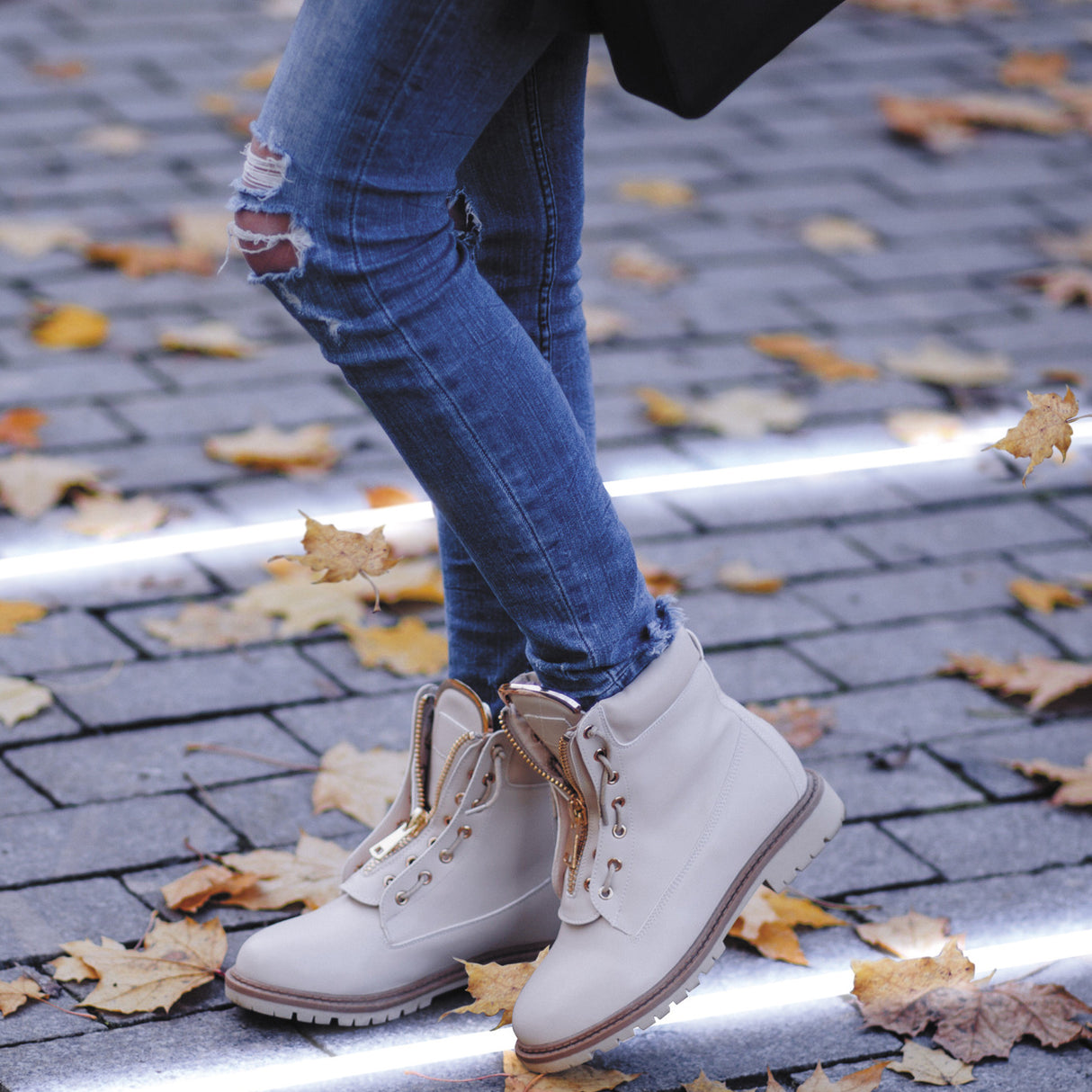 A person stands on a cobblestone path with autumn leaves, illuminated by the Drive-safe Embedding LED Profile ~ Model Dublin XL, which glows as a white line along the ground. They wear light beige boots and ripped blue jeans.