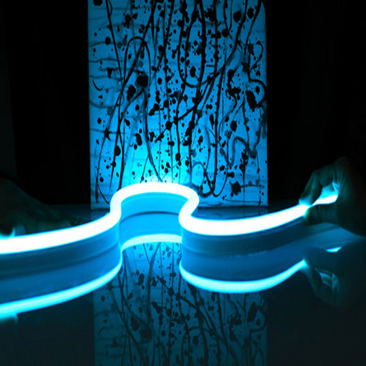 Two hands hold the Flexible LED Neon Light Silicone Profile ~ Model Neon Las Vegas, a glowing blue squiggle, in front of a splatter-painted canvas with its reflection visible on a shiny surface—ideal for creative decorative lighting.