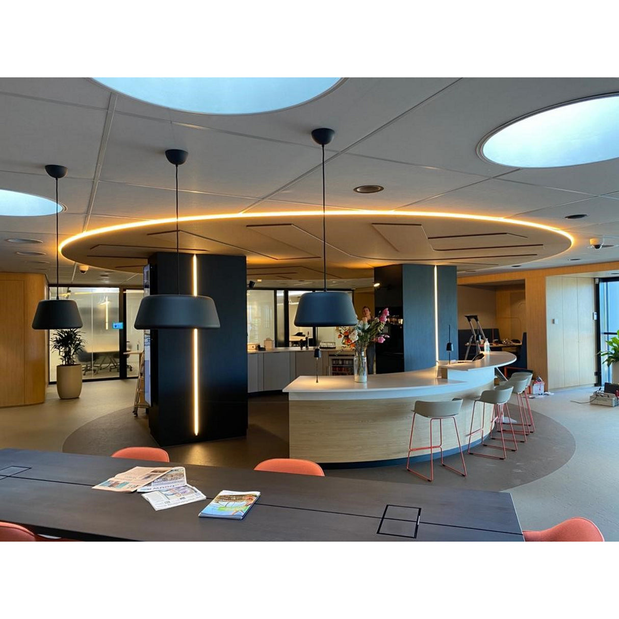 Modern office kitchen with a curved island, high chairs, and pendant lights accented by the Ultra Thin Bendable LED Strip Channel ~ Model Milano Slim. A table with magazines is in the foreground; plants and glass-walled offices are in the background.