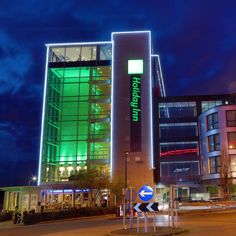 A modern Holiday Inn hotel at night is illuminated by green and white Hi-Lite 30 Straight Section Colored Border Contour Tube Lights. Large glass windows, the hotel’s logo, cars, and a roundabout are visible in the foreground.