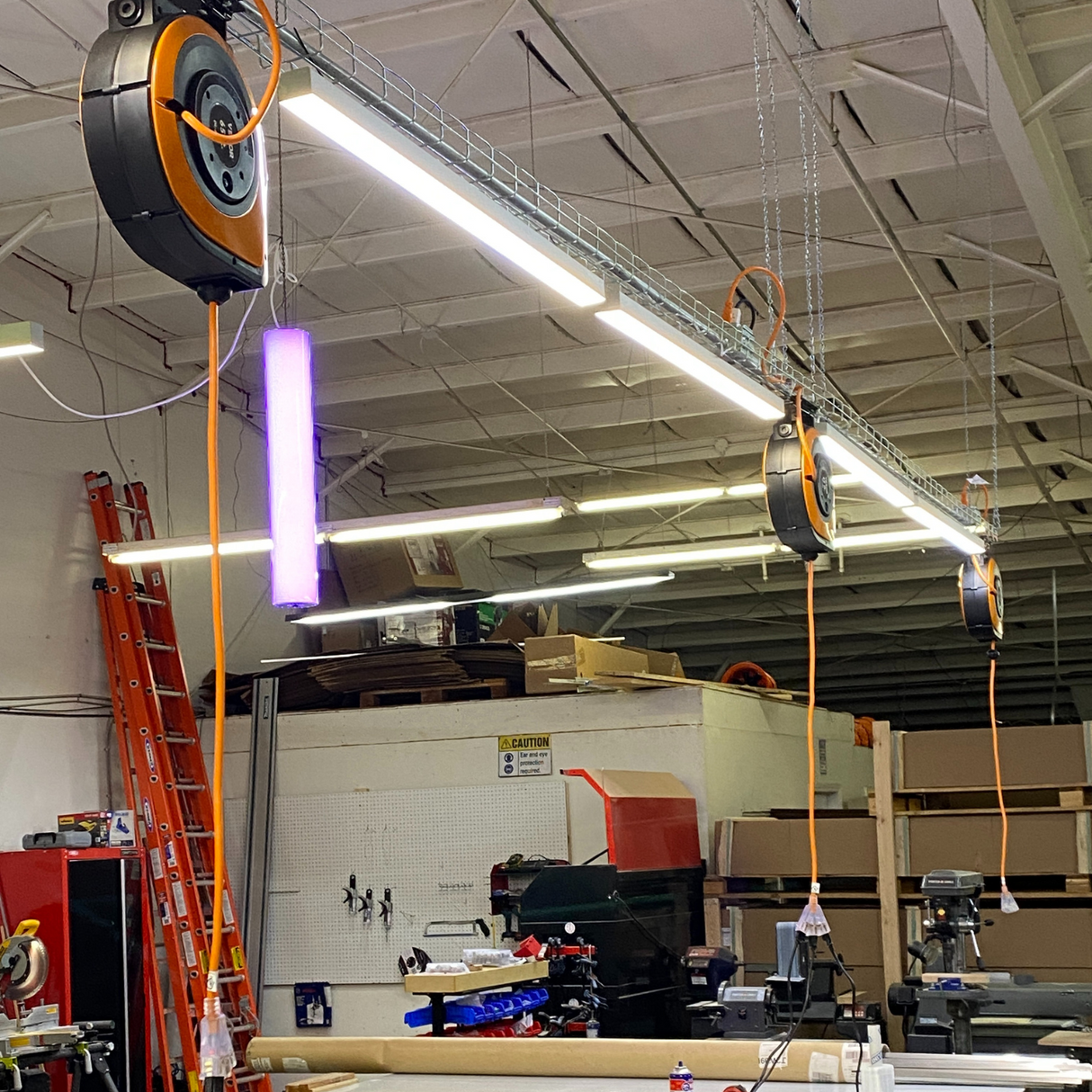 A workshop features retractable extension cords, fluorescent lights with diffusers, tools and equipment. Cardboard boxes, a ladder, and a 2 Wide Aluminum Linear LED Lighting Profile ~ Model PL55-FL are visible in the background.