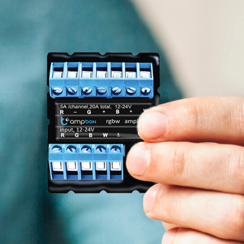 A hand holds the ampBox by BleBox, a 20A 4-Channel LED amplifier/repeater with blue and silver screw terminals for connecting wires in RGBW lighting setups, labeled input, 12-24V.