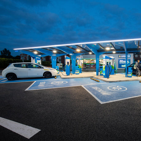 A white electric car charges at dusk at a modern EV station, illuminated by Colored Border Contour Tube Light ~ Hi-Lite 30 Straight Section. The station has multiple charging points, blue signage, and a small convenience store in the background.