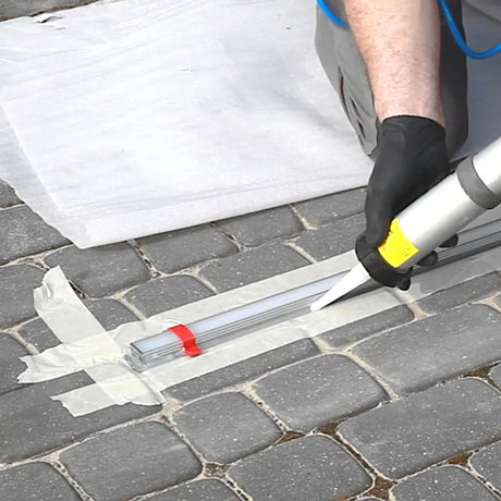A person uses black gloves to apply sealant with a caulking gun between cobblestones, using tape and the Installation Guide Clip for HR-MAX Profile to achieve a neat line—ideal for HR-MAX profile system or lighting installation videos.