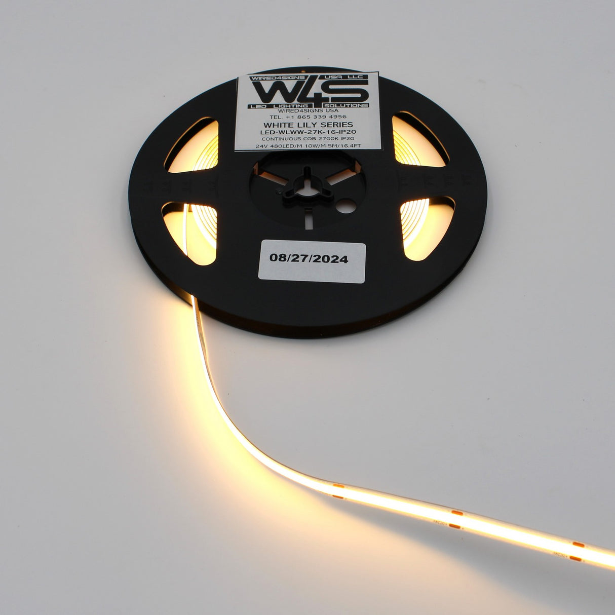 A black spool holds a Continuous COB IP20 LED Strip (24V) ~ White Lily Series, partially unrolled and glowing. Labels display product details and the date 08/27/2024, ideal for decorative lighting projects.