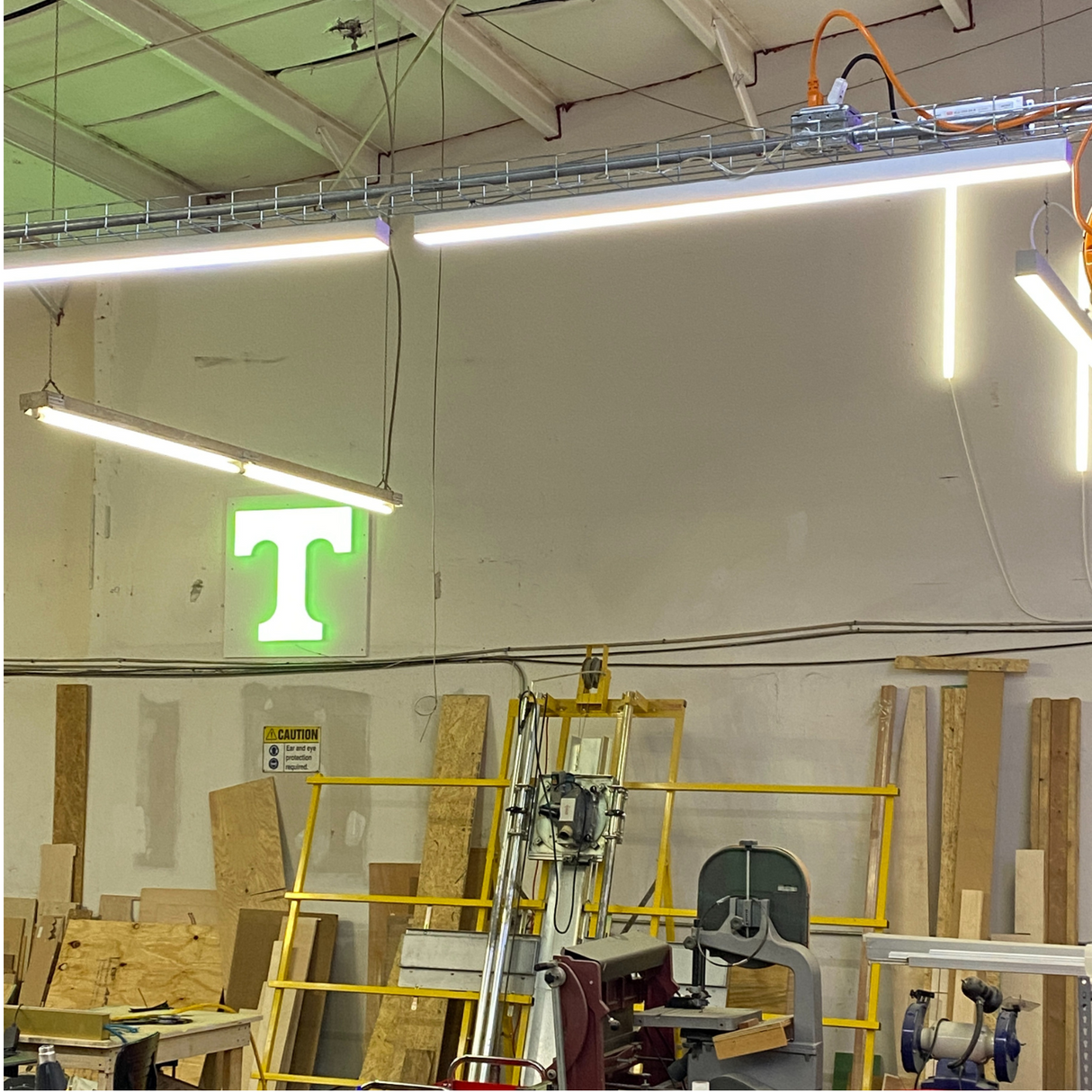 A workshop with wooden planks, tools, a yellow ladder, and fluorescent lights. 2 Wide Aluminum Linear LED Lighting Profile (Model PL55-FL) with diffuser options hangs from the ceiling; a glowing green T sign is on the wall.