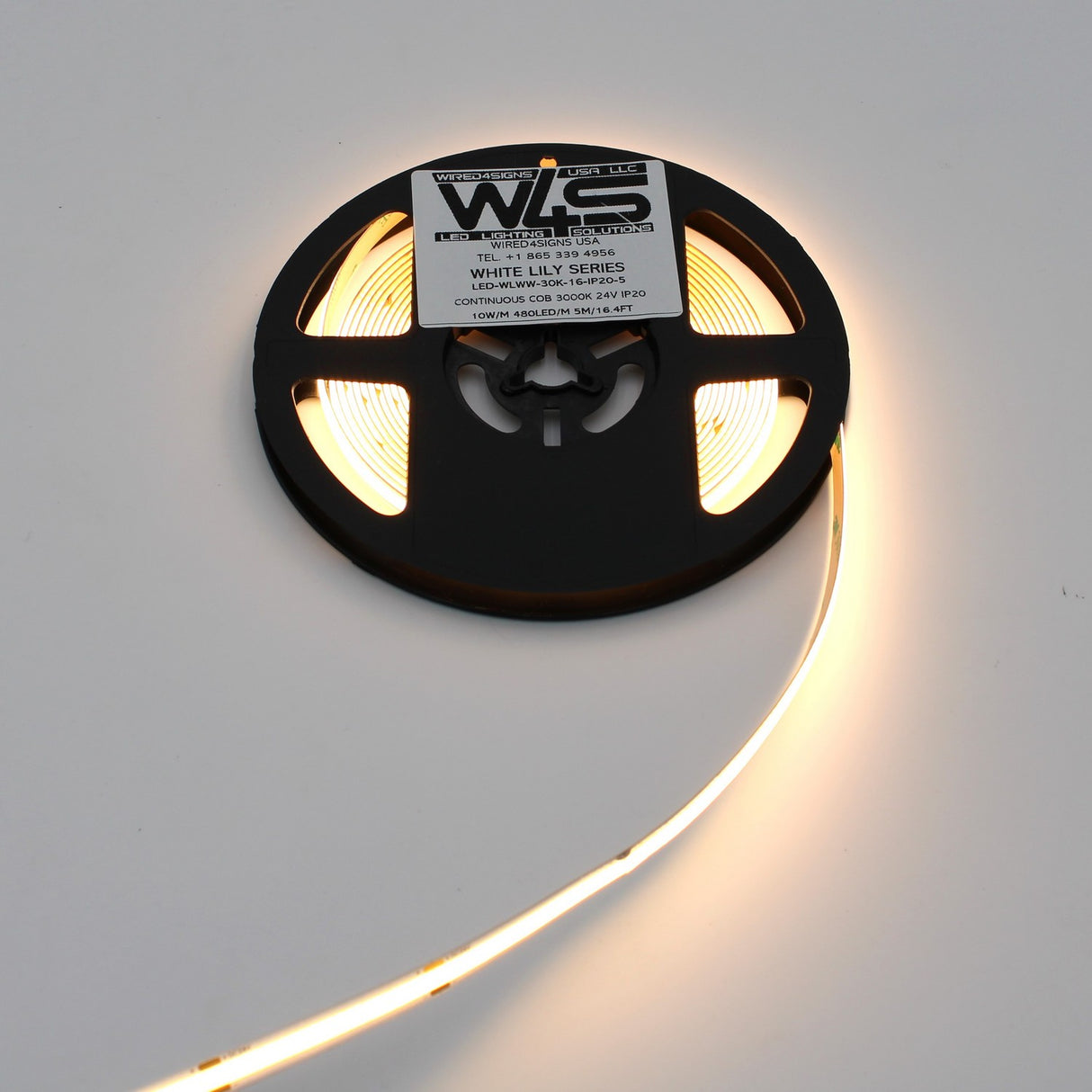 A spool of Continuous COB IP20 LED Strip (24V) ~ White Lily Series glows softly when partially unrolled on a white surface. The black reel’s label shows product details, ideal for continuous and decorative lighting applications.