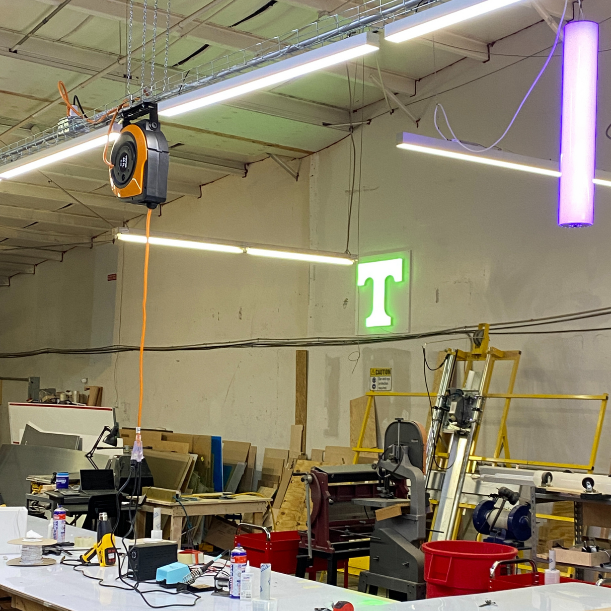 A workshop filled with tools and materials has a glowing green T sign on the back wall, bright overhead light from a 2 Wide Aluminum Linear LED Lighting Profile (Model PL55-FL), and a purple ceiling fixture.