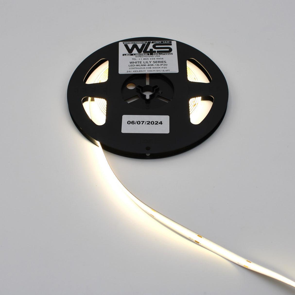 A reel of Continuous COB IP20 LED Strip (24V) ~ White Lily Series, partially unspooled and glowing with seamless light; a label on the reel shows product details and date 06/07/2024.