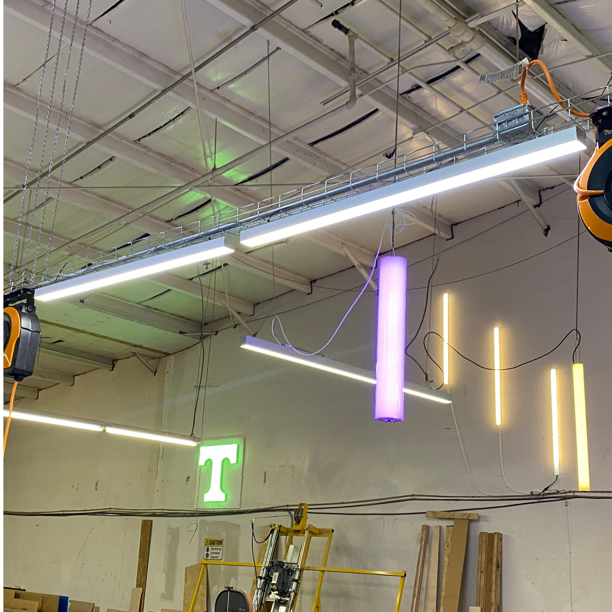 A workshop ceiling with fluorescent lights, a hanging purple cylindrical light, PL55-FL 2 wide aluminum linear LED lighting profiles on the wall, a green illuminated T sign, and various construction materials below.