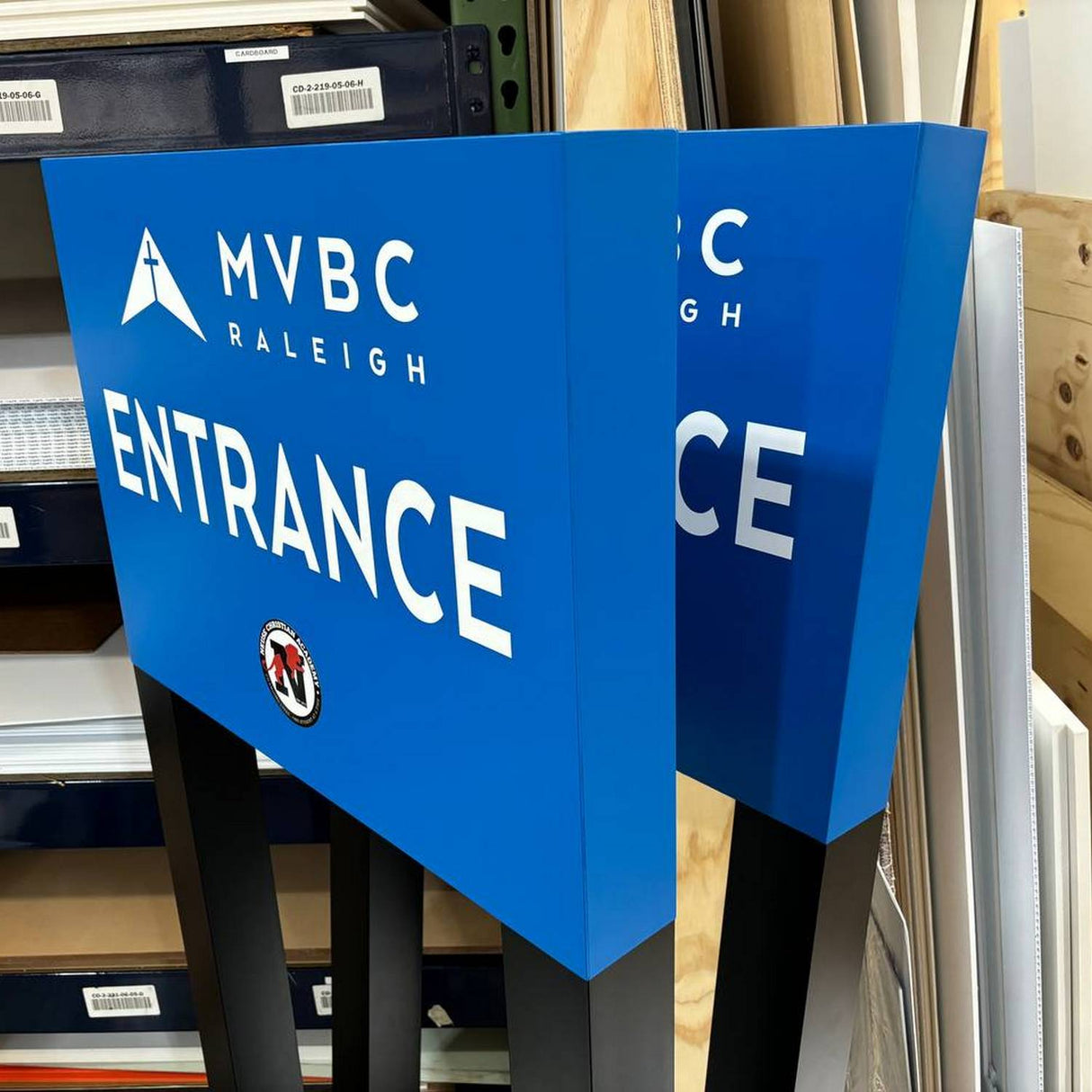 A blue double-sided sign with MVBC Raleigh ENTRANCE in white and a logo below stands on black posts in a workshop with shelves of industrial products, including Black 2-part Methacrylate Adhesive ~ W4S 55300 (400ml 1:1 Mix / C-System).