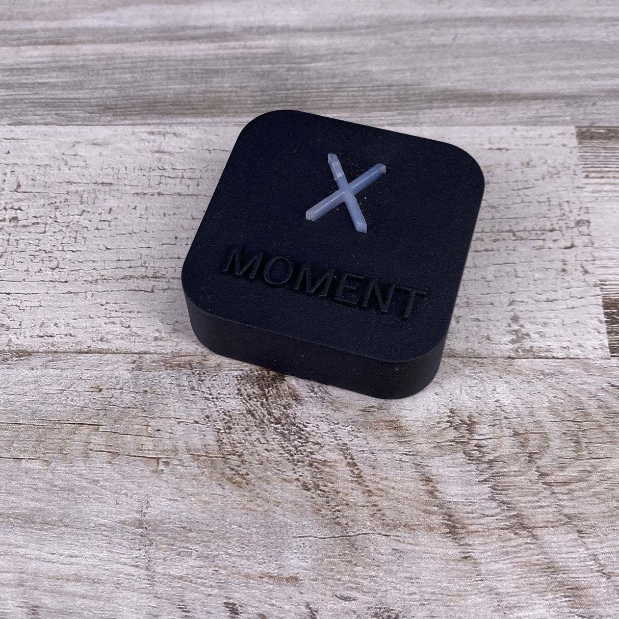 A compact black, square device with a white “X” and “MOMENT” engraving rests on light wood—perfect as the Casambi Lighting Controller with Alexa Voice Control, Estol X-Moment.