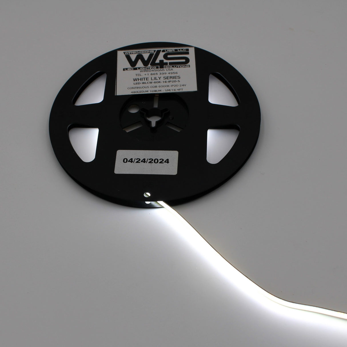 A black reel labeled Continuous COB IP20 LED Strip (24V) ~ White Lily Series dispenses a glowing white strip for seamless lighting, with a sticker showing the date 04/24/2024 also visible on the reel.