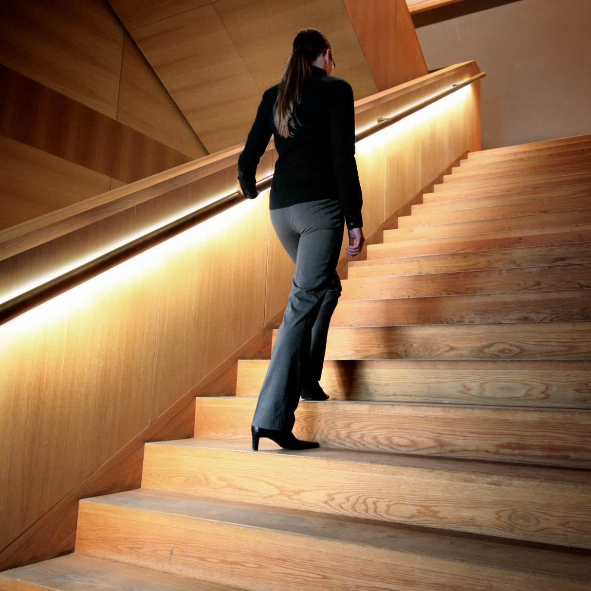 A woman in business attire and high heels ascends a modern wooden staircase featuring Round Stainless Steel LED Handrail Profile ~ Model Comenza in a well-lit, contemporary interior.