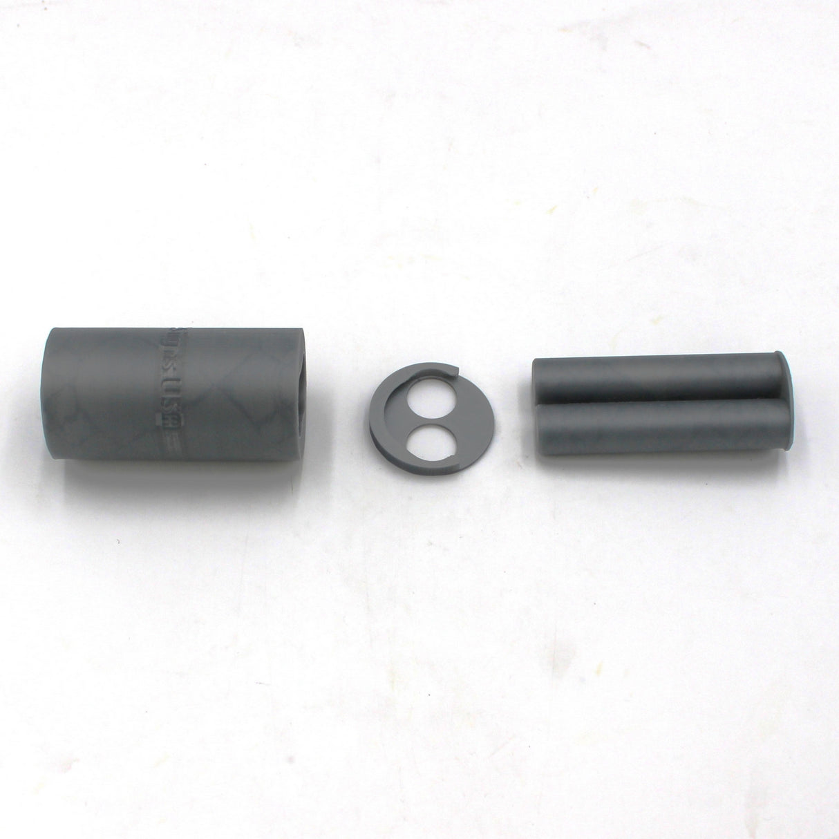 Three cylindrical black objects and one round flat black piece with two holes, seen in a row on a plain white surface, are parts of the 3D-Printed Caulking Gun Adapter for Methacrylate 50ml 1:1 Mix.