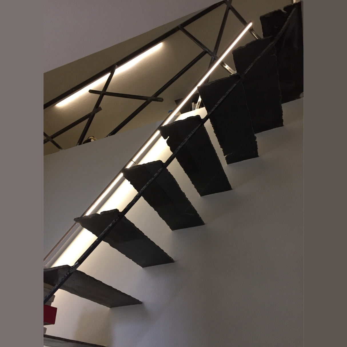 A modern staircase with black metal steps and a Round Stainless Steel LED Handrail Profile ~ Model Comenza, viewed from below against a white wall, features exposed ceiling beams and a linear light above.