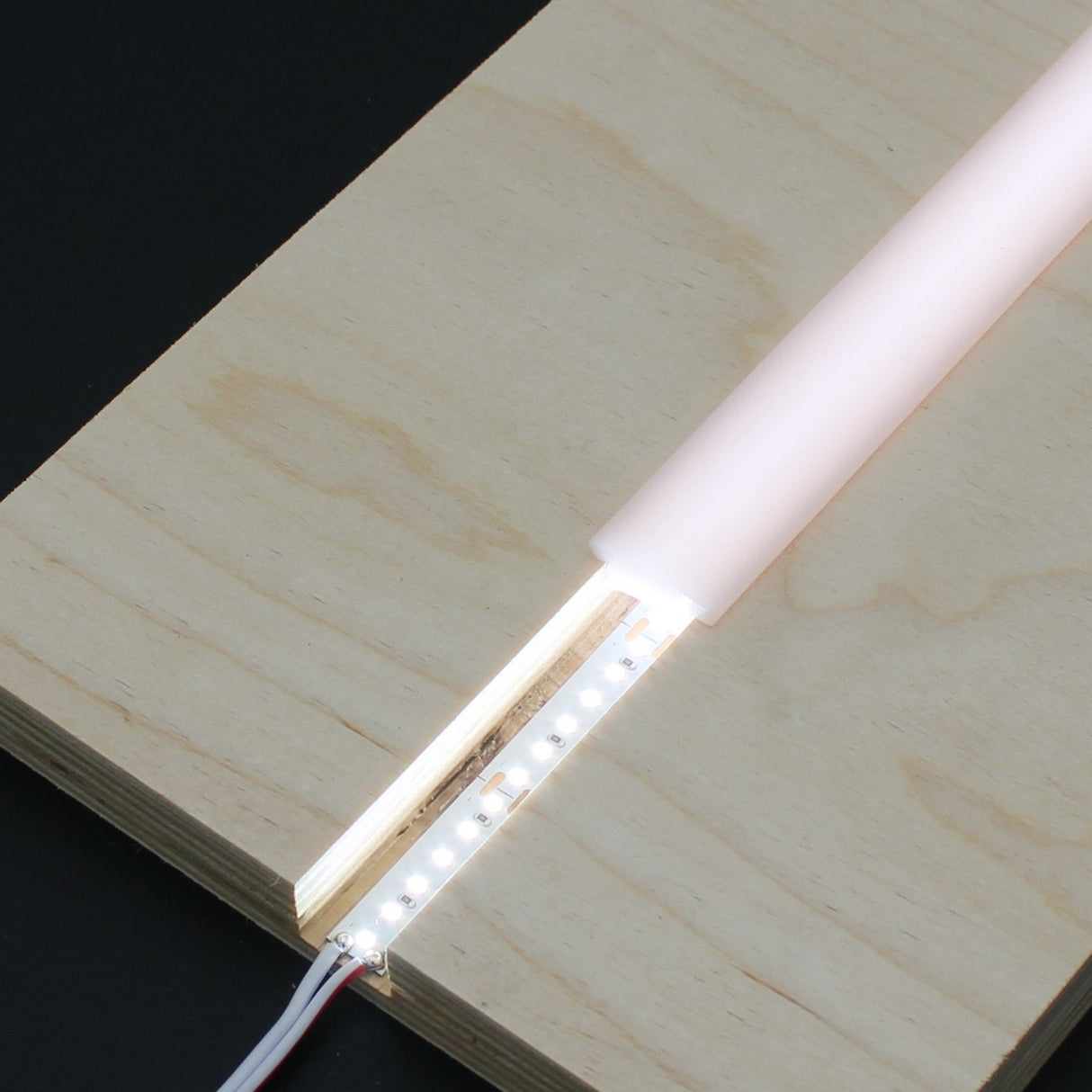 A close-up of a light fixture featuring the Arles LED Strip Flexible Diffuser embedded in a wooden board and partially covered by a frosted plastic diffuser, casting a gentle white glow.