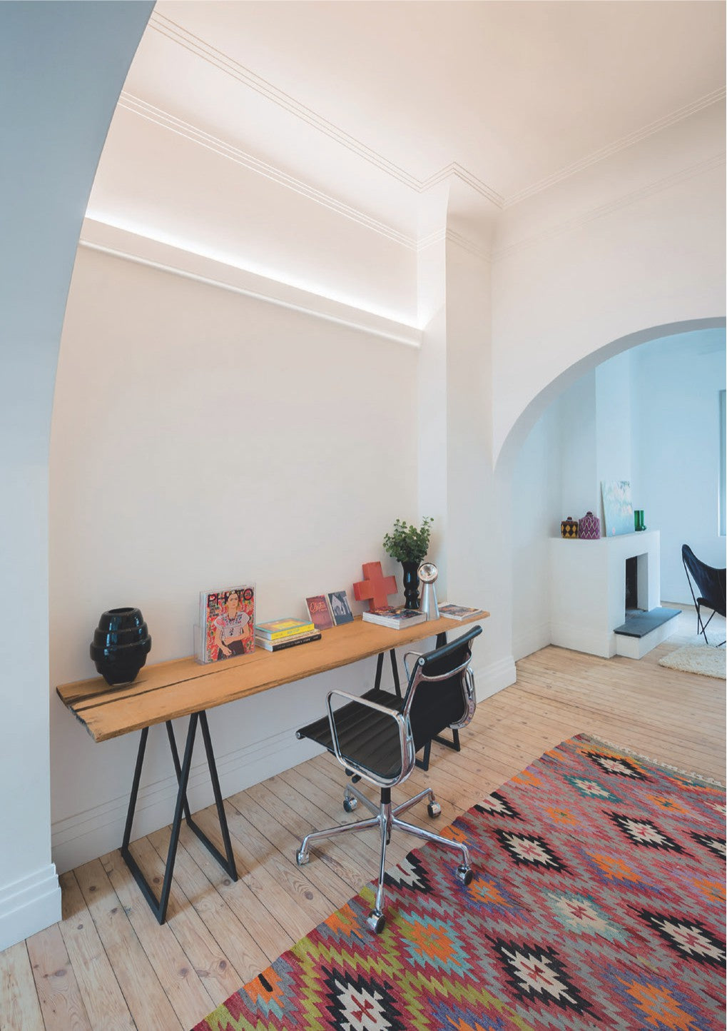 A modern home office features a wooden desk, black chair, colorful magazines, vibrant geometric rug, and light wood floors. The Up and Down Ceiling Wall Wash LED Profile ~ Model Badalona provides balanced illumination for versatile lighting applications.