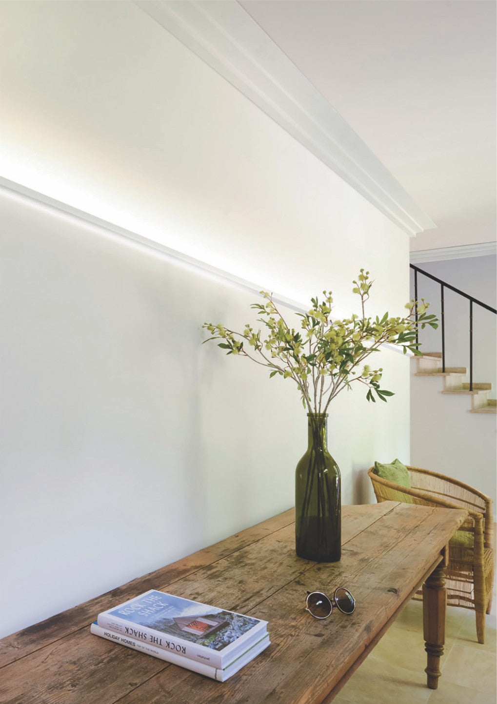 A green glass vase with leafy branches rests on a wooden table beside sunglasses and books; balanced illumination from the Up and Down Ceiling Wall Wash LED Profile ~ Model Badalona enhances the bright, minimalist room with wicker chair and staircase.