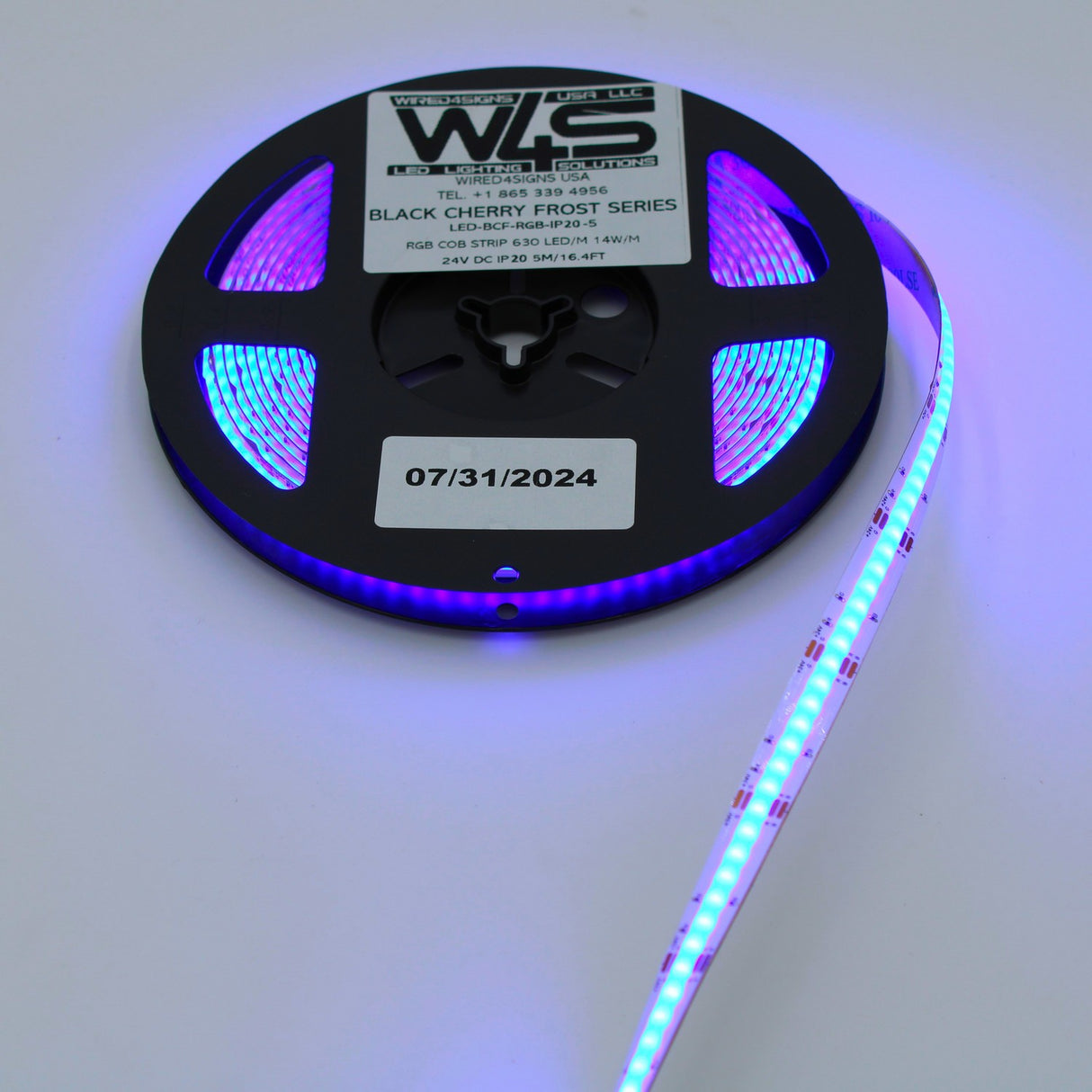 A spool of RGB COB IP20 LED Strip (24V) ~ Black Cherry Frost Series glows purple and blue. A label dated 07/31/2024 is attached to the black reel.