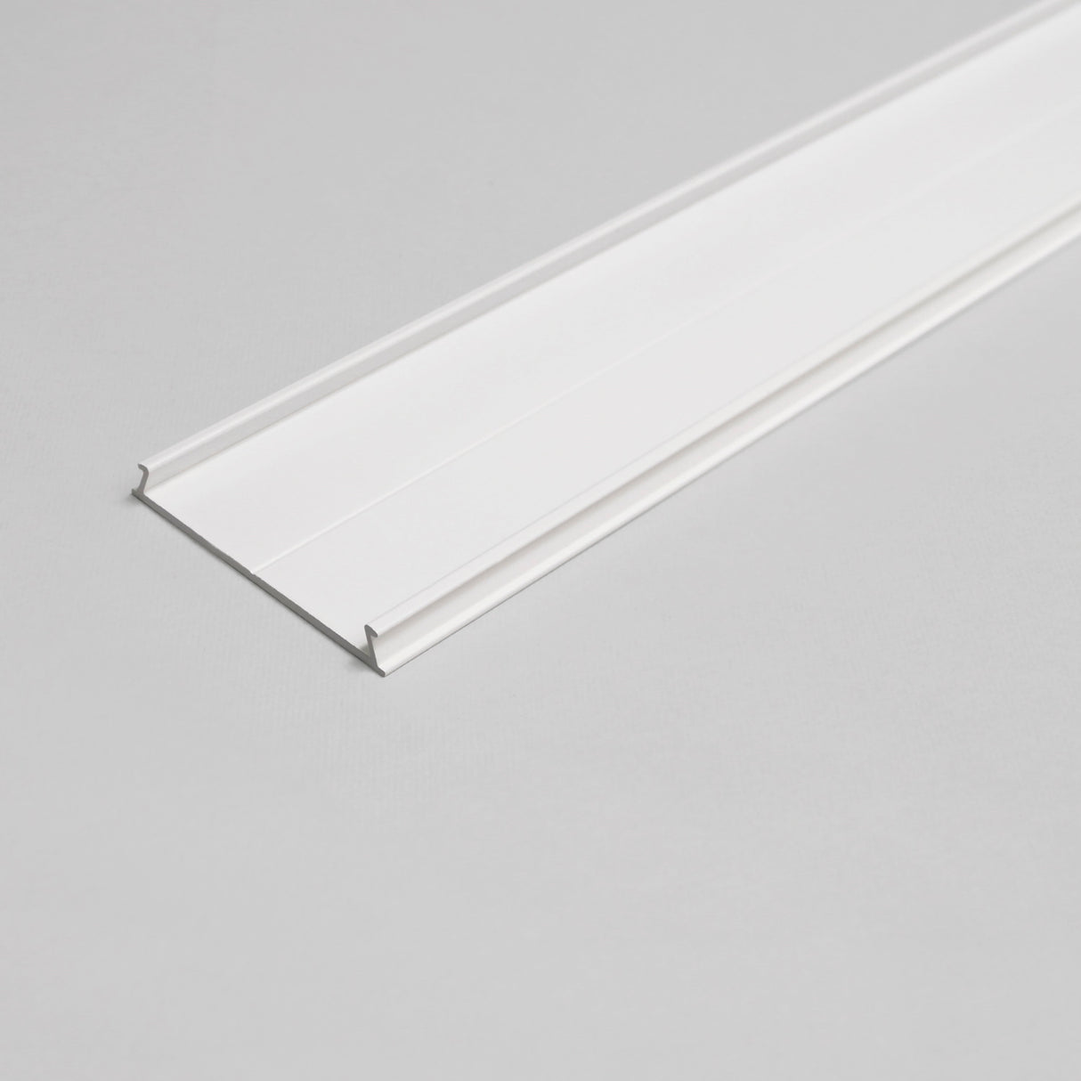 The Complementary Cover Profile ~ Model C10, a white, flat, rectangular trim strip with slightly raised edges ideal for LED profiles, is shown on a light gray surface in an angled overhead photo.
