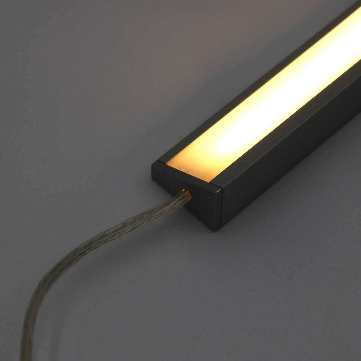 A close-up of the White Universal Surface-mount LED Corner Lighting Fixture (24V) ~ Corner14 Series, featuring a warm white glow in a dark metal frame with a transparent power cord, ideal for under cabinet lighting against a plain background.