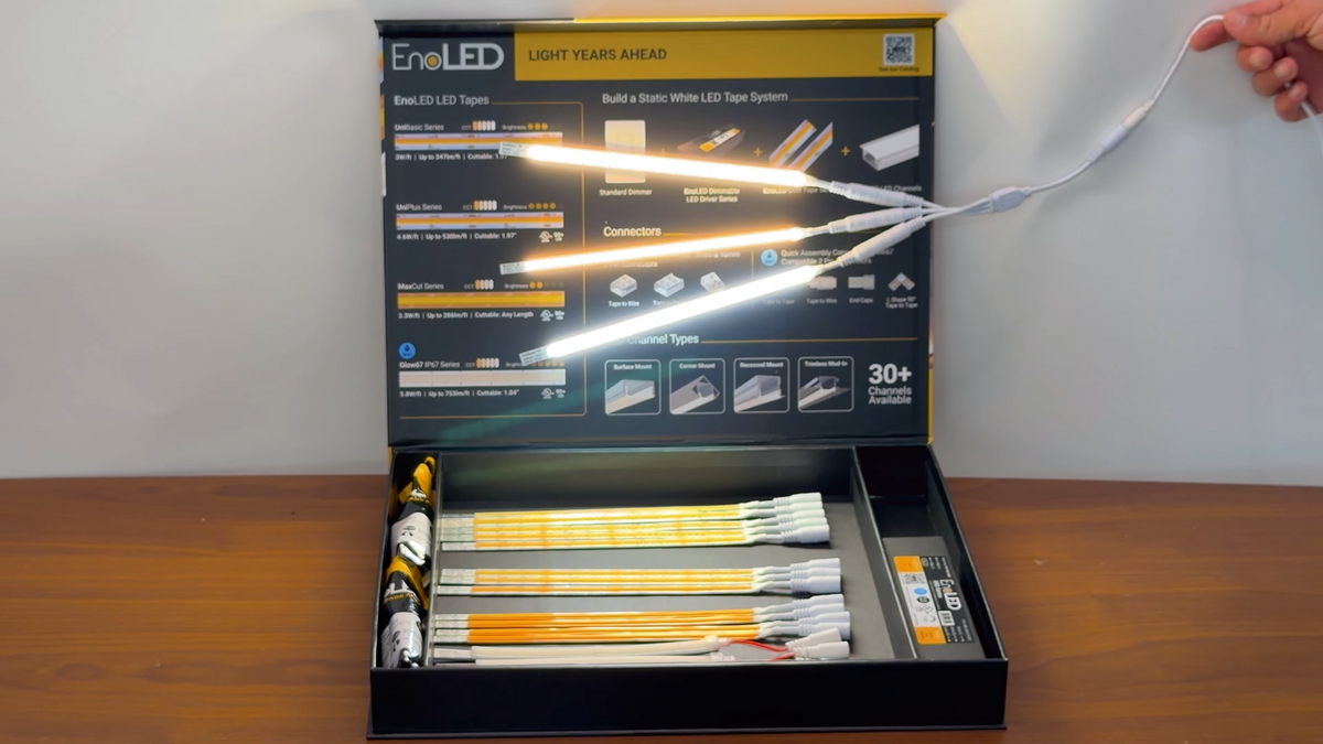 A hand holds glowing LED tape samples above an open EnoLED LED Strip Sample Box, which displays various neatly arranged LED components on a table.