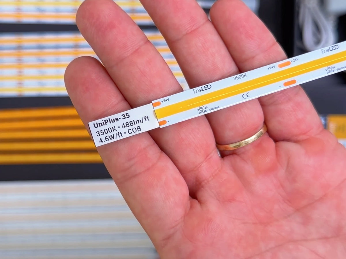 A hand holds an LED strip labeled UniPlus-35, 3500K, 488 lm/ft, 4.6 W/ft, COB from the EnoLED LED Strip Sample Box. Other LED tape samples are blurred in the background.