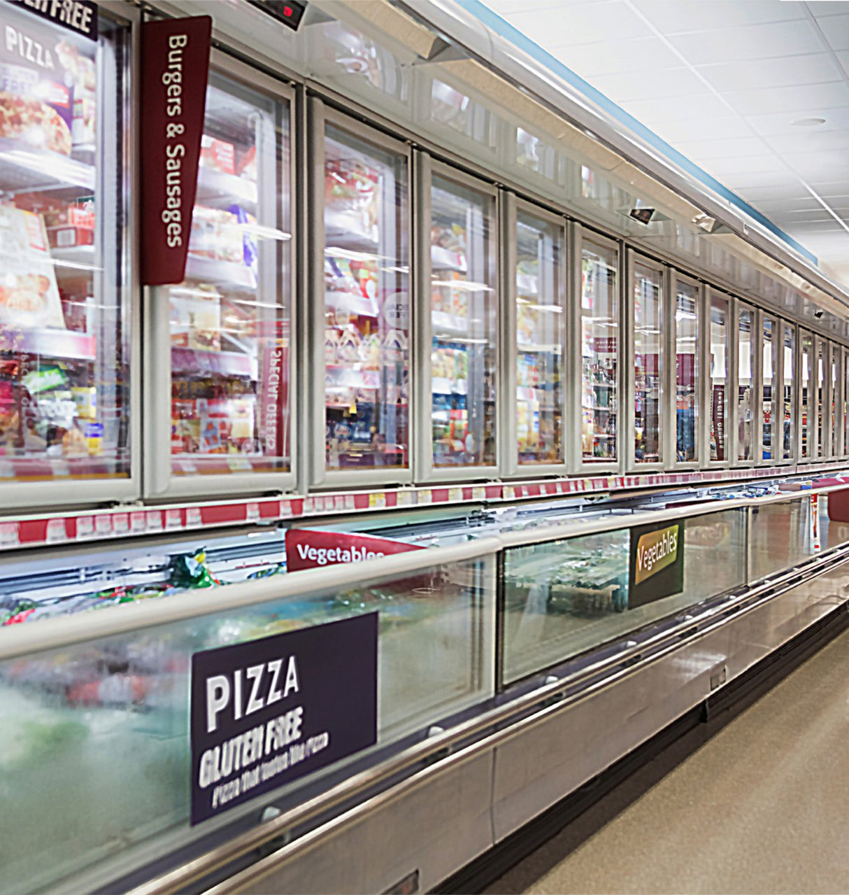 A supermarket frozen food aisle features glass-door freezers lit by the Commercial Refrigerator Lighting LED System ~ FLX Stix CDV Series, brightly illuminating signs for burgers, sausages, vegetables, and gluten-free pizza.