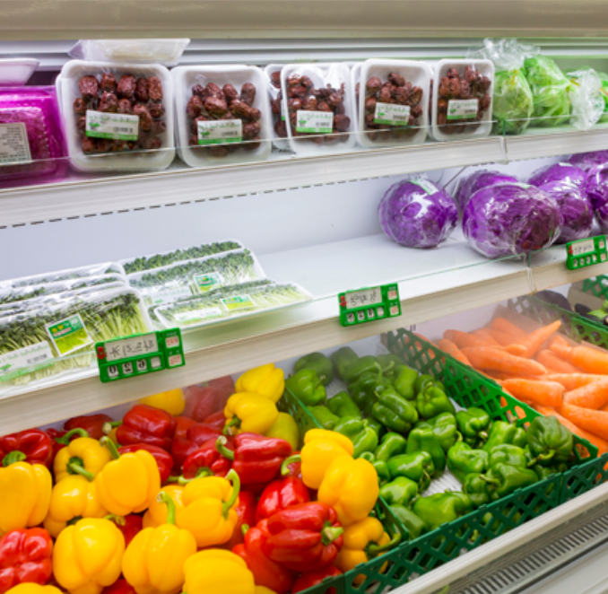 A supermarket produce section, lit by the FLX Stix CDV Series Commercial Refrigerator Lighting LED System, displays packaged vegetables above and loose yellow, red, and green bell peppers, green peppers, carrots, and cabbage below.