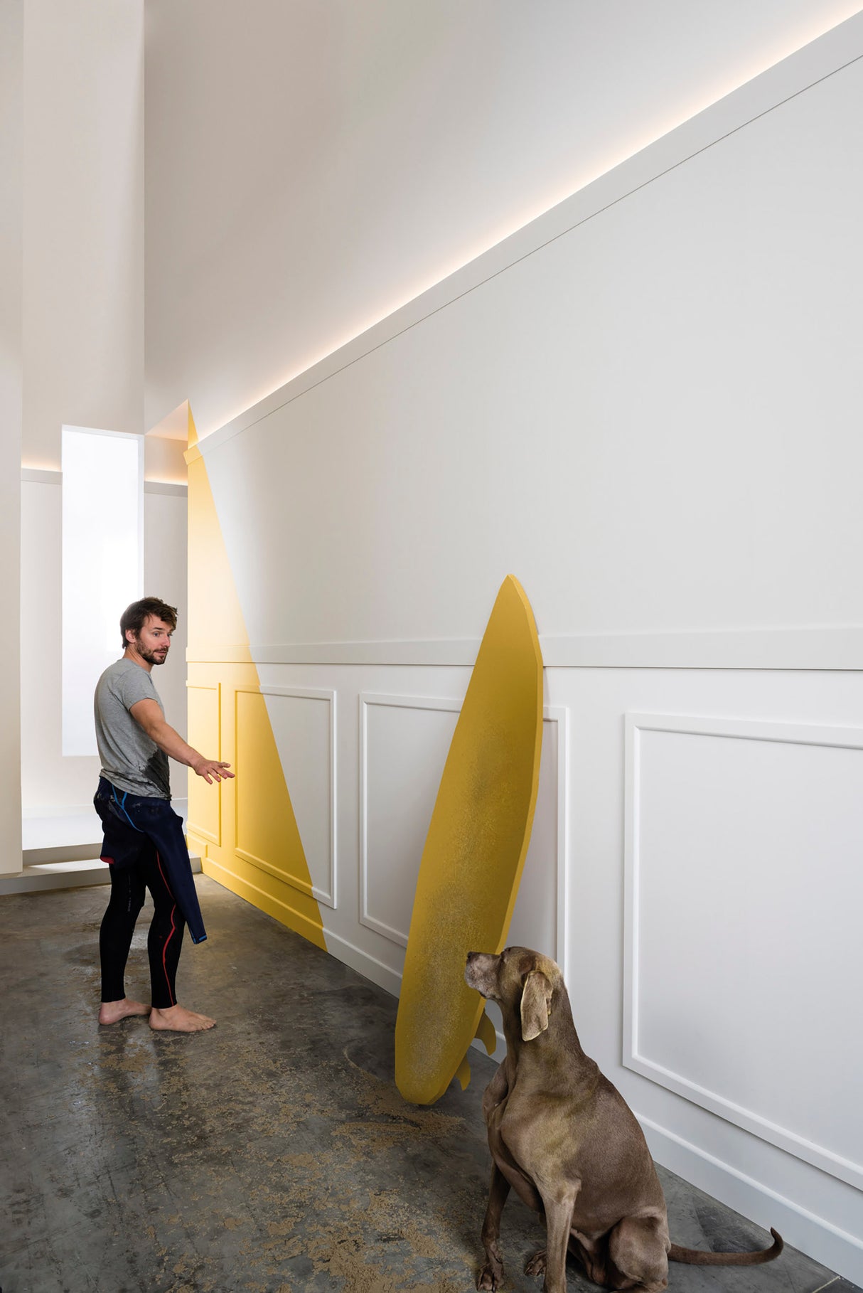 A man gestures toward the Indirect Ceiling Wall Wash LED Profile ~ Model Galicia illuminating a large yellow translucent shape leaning against a white paneled wall, while a brown dog sits on the concrete floor in the modern, brightly lit interior.