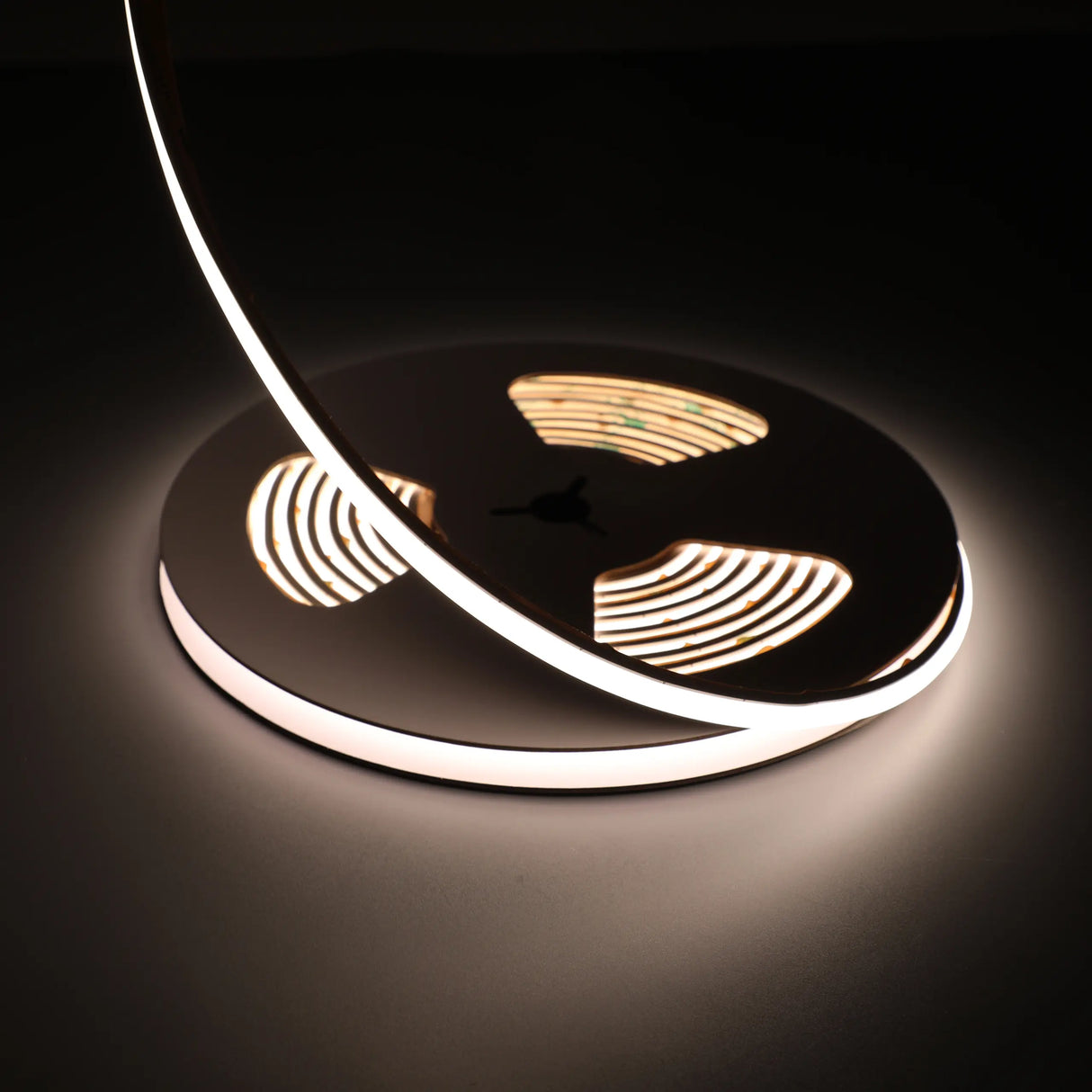 A close-up of the Flexible Dot-less High Power COB IP67 LED Strip (24V) ~ Glow67 Pro Series, wound in a circle and softly glowing in the dark, displays high-performance lighting with smooth reflections and shadows on the surface below.