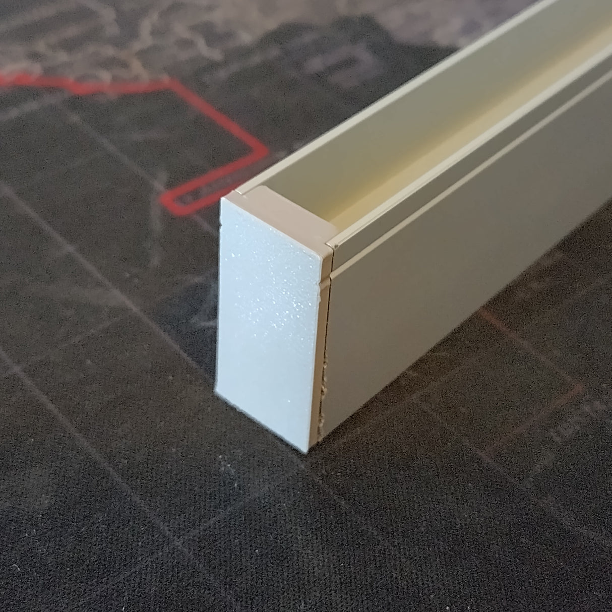 A close-up of a rectangular aluminum Hannover Profile featuring the Plastic End Cap, resting on a dark surface with red and gray patterns.