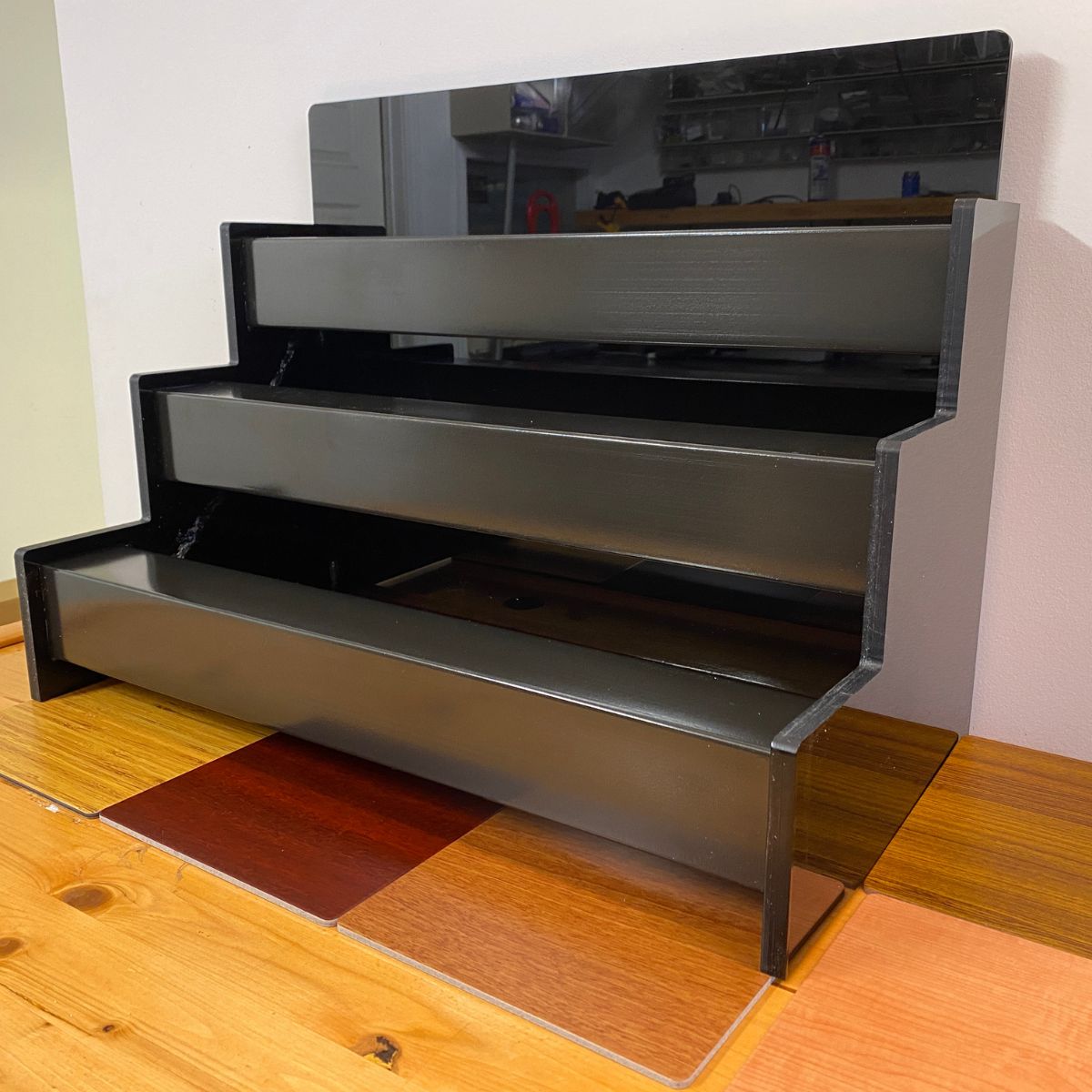 A three-tiered black display stand with a mirrored back sits empty on a checkered wooden table, against a light-colored wall—perfect for showcasing the 2.91 Square Polycarbonate LED Lighting Tube ~ Model Holston74.