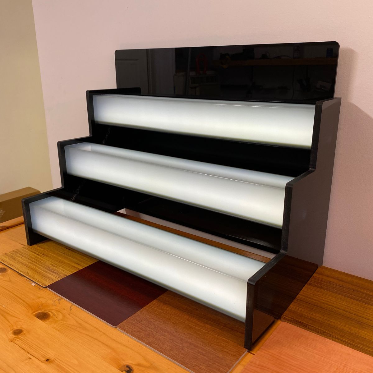 A three-tiered black display shelf features sleek 2.91 Square Polycarbonate LED Lighting Tubes (Model Holston74) with white illuminated steps. The minimalist unit sits empty on a wooden surface against a beige wall, ready for display.