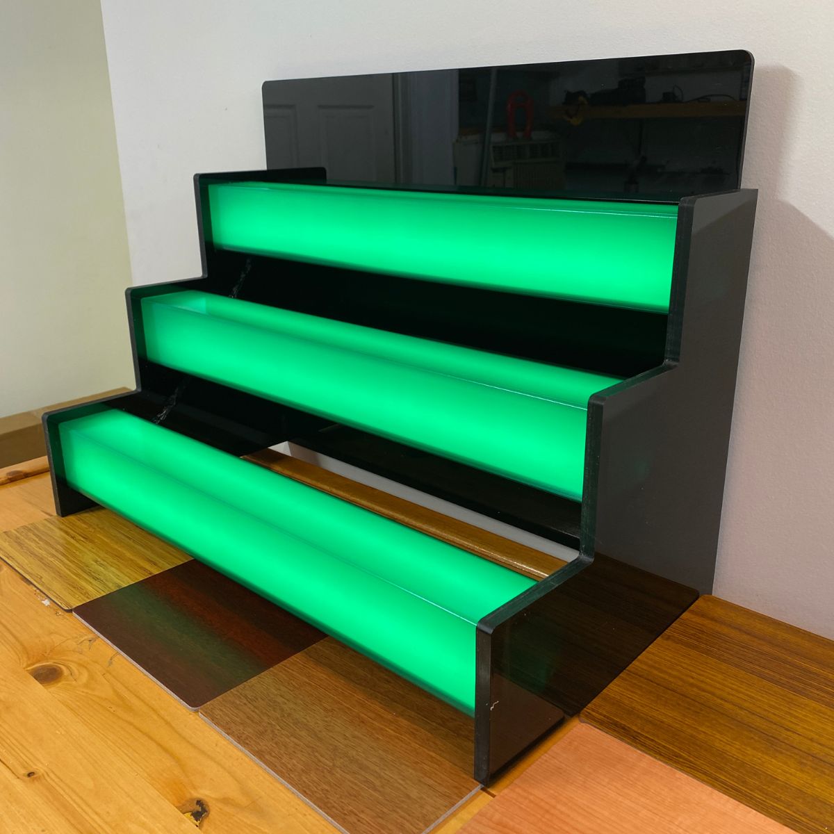 The 2.91 Square Polycarbonate LED Lighting Tube ~ Model Holston74 is a three-tiered black display stand with glowing green shelves, each featuring integrated RGB pixel LEDs for illuminated, staircase-like steps on a wooden surface against a white wall.