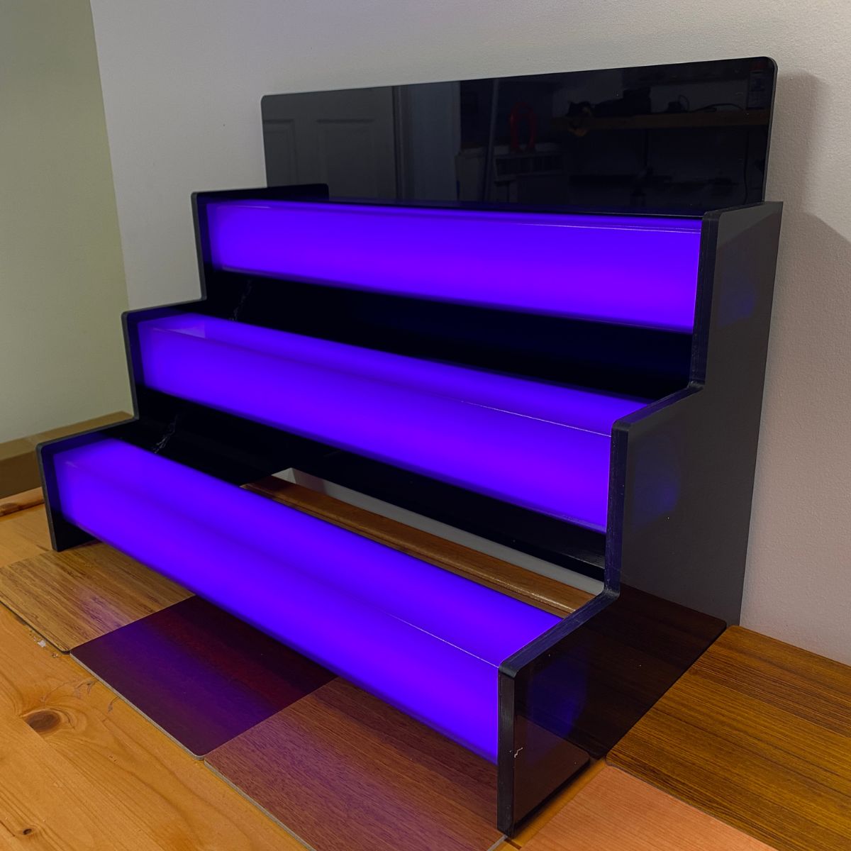 A black, three-tiered display stand features 2.91 Square Polycarbonate LED Lighting Tubes (Model Holston74) glowing purple beneath each tier, set on a wooden surface against a light-colored wall.