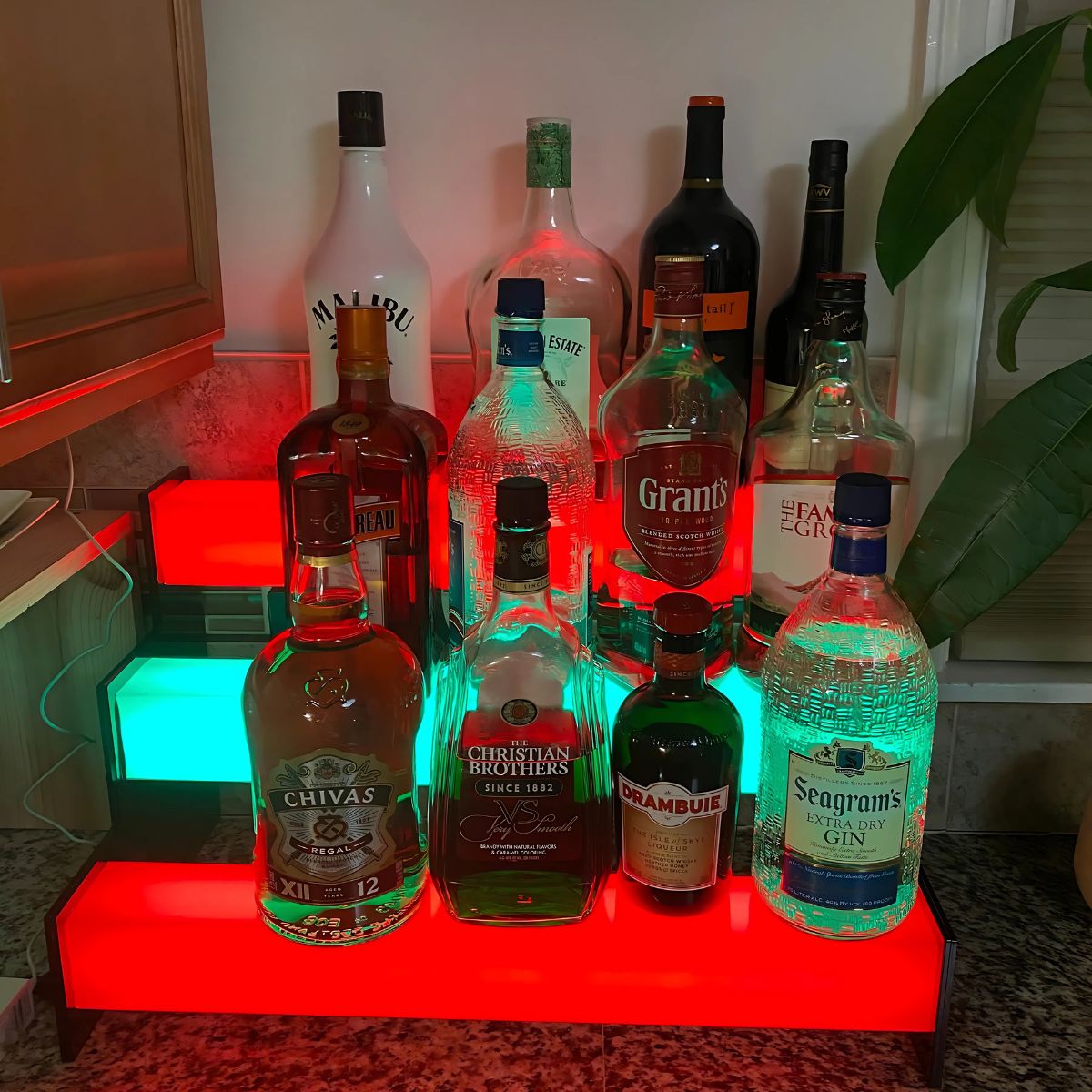 A variety of liquor bottles are displayed on red and green shelves with an RGB pixel LED strip using the 2.91 Square Polycarbonate LED Lighting Tube ~ Model Holston74, set on a kitchen countertop. A large plant sits to the right.