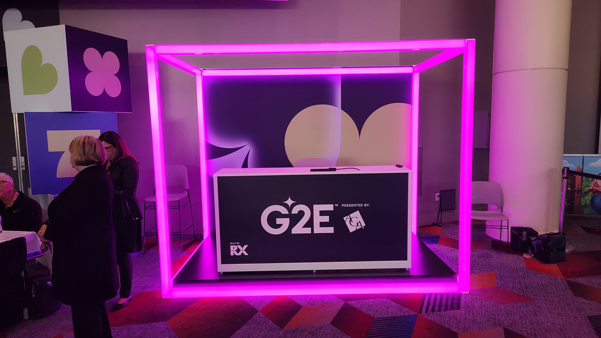A booth with a G2E sign is framed by a glowing pink neon cube made from 2.91" Square Polycarbonate LED Lighting Tube ~ Model Holston74. People stand nearby on colorful patterned carpet with geometric decorations in the background.