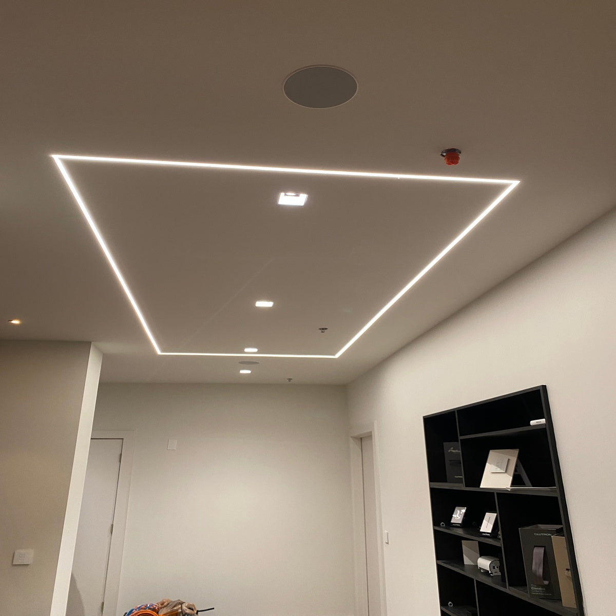 A modern ceiling features recessed square lights and a border of 0.39 Plasterboard Trimless LED Channel (Model Hide10), creating a sleek, continuous line of light that frames the minimalist room with its white walls and shelving.