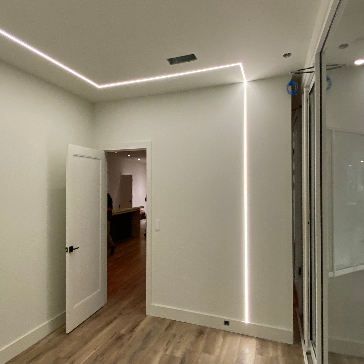 A modern room with light wood floors, white walls, and an open white door features a continuous line of light from the 0.39 Plasterboard Trimless LED Channel (Model Hide10) embedded in the ceiling and wall. A glass wall is visible on the right side.