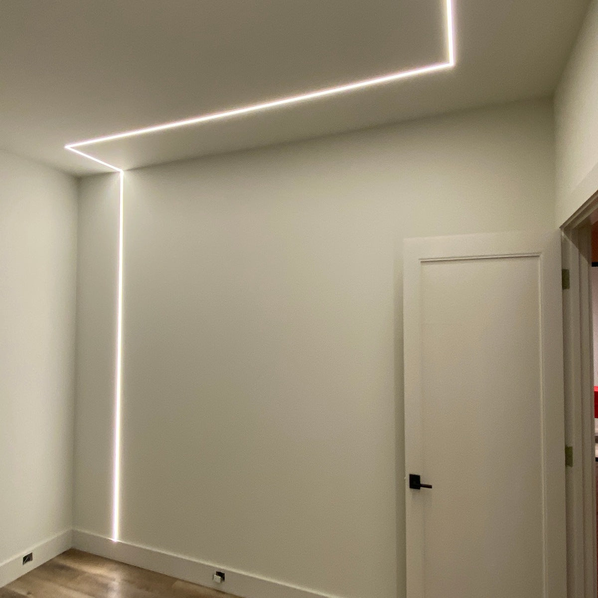 A minimalist room with white walls and a partially open white door features the 0.39 Plasterboard Trimless LED Channel (Model Hide10), creating a continuous line of light along the ceiling and down one wall.