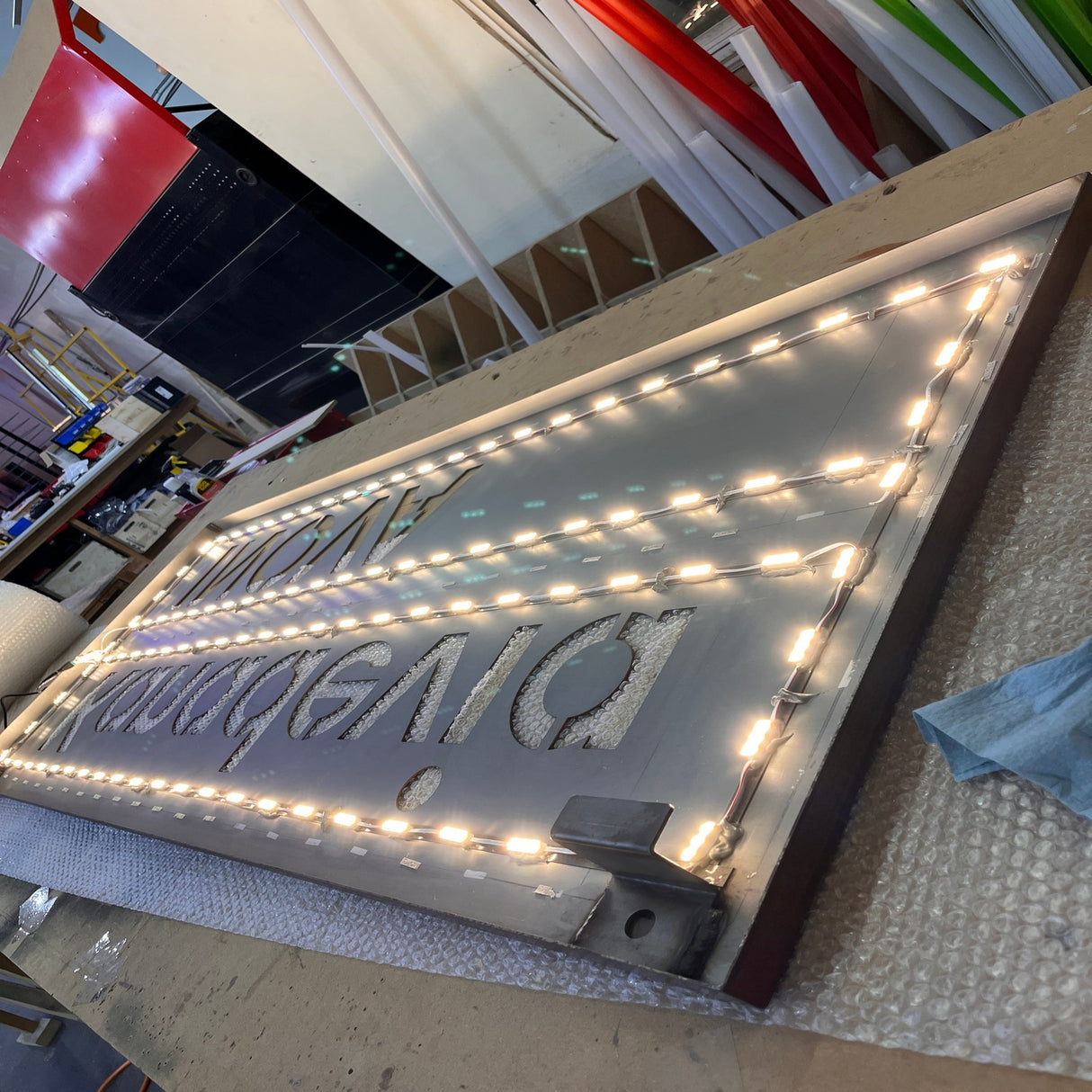 A rectangular sign featuring the Mini3 White 160 Mini LED Module ~ Wide Angle Series outlines reversed text and lies flat on a worktable in a workshop, surrounded by tools, materials, and rolls of colored film.