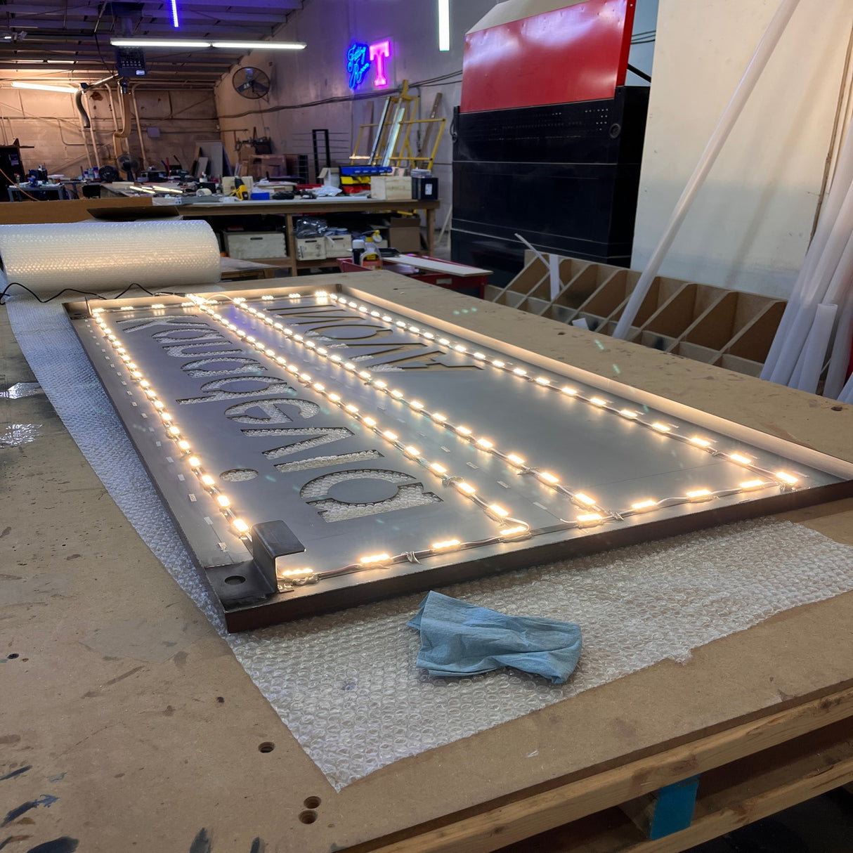 A large metal sign with cut-out letters and Mini3 White 160 Mini LED Module ~ Wide Angle Series strips lies flat on a workbench in a workshop, surrounded by tools, materials, and neon signs; photo taken with a wide-angle lens.