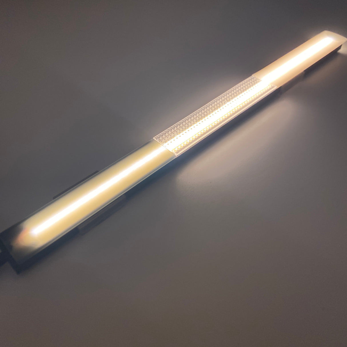 The Ultra Thin Bendable LED Strip Channel ~ Model Milano Slim emits a warm white glow, illuminating a smooth, light-colored surface and casting soft, subtle shadows.