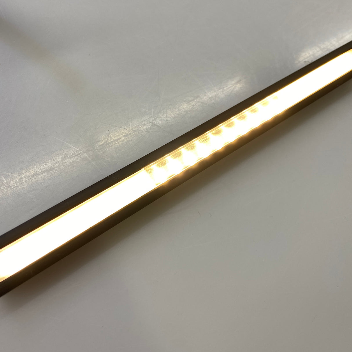 A close-up of the slim LED Channel Cover ~ C2 Click, fitted on a smooth off-white surface. It emits a warm white glow, with individual bulbs partially visible through its sleek diffuser cover.