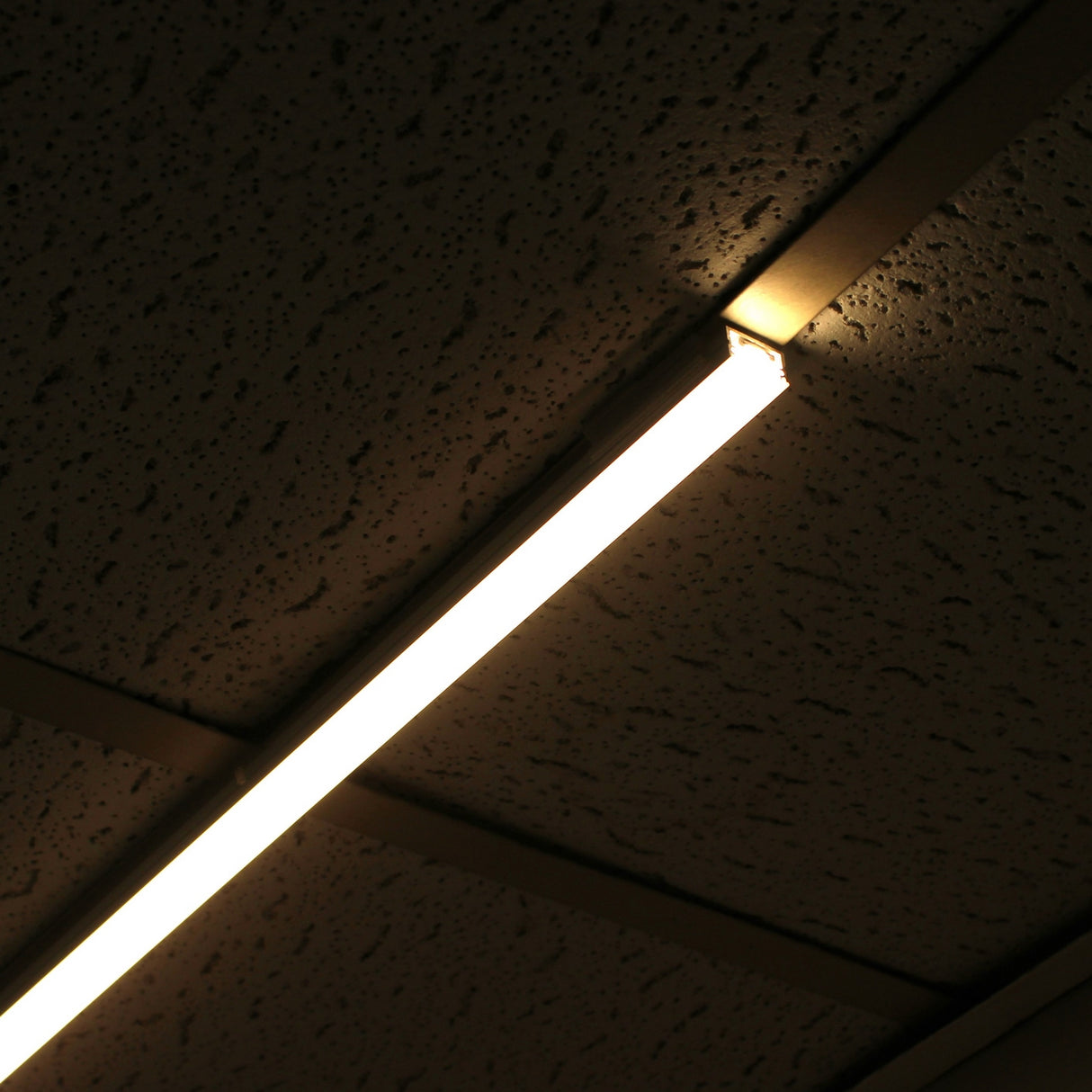A Roma XL Drop Ceiling Clip secures a fluorescent light fixture to a speckled acoustic ceiling, illuminating a dimly lit room. The photo is shot from below at an angle, highlighting the sturdy 3D printed clip in use.