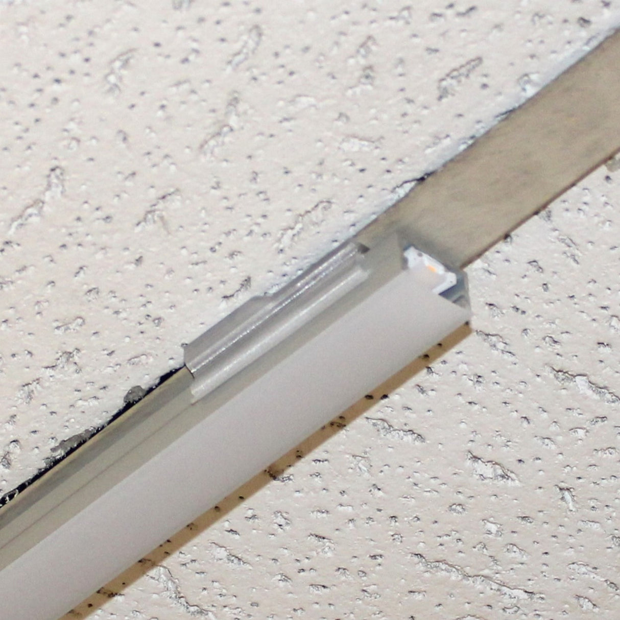 Close-up of a Roma XL Drop Ceiling Clip securing a recessed fluorescent light fixture in a white textured drop ceiling with visible metal support grid and frame.