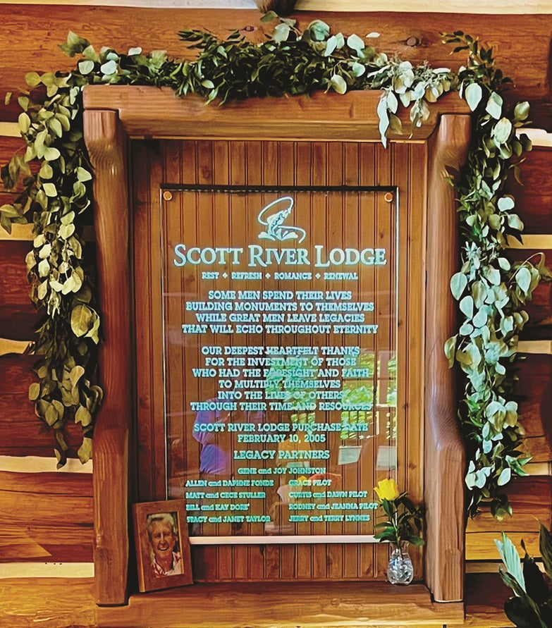A wooden Scott River Lodge plaque with white and blue text, decorated with a green garland, rests on a 1/4 LED Edge-lit Acrylic Sign Base Channel ~ Model Mikro10, next to two photos, a yellow rose in a vase, and two small stones.